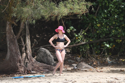 Emily Blunt in bikini posing near tree and rocks, wearing black swimsuit and bright pink wide-brimmed hat