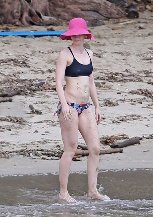 Emily Blunt in bikini walking along shoreline, wearing black bikini top and floral bikini bottoms with pink sun hat