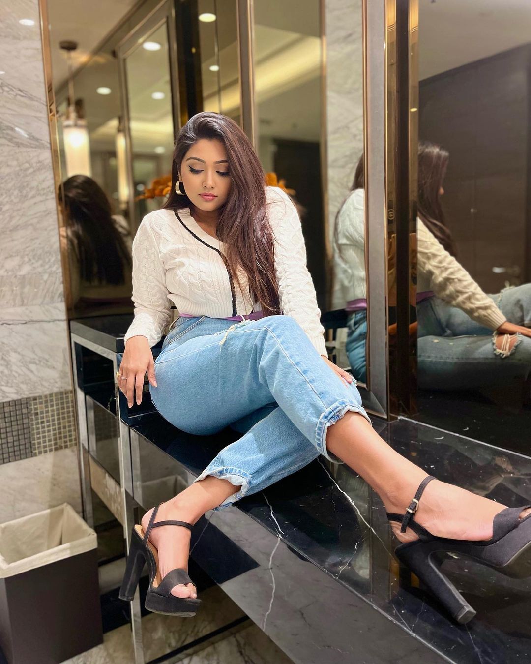 Agnijita Banerjee looking down and sitting on a black slab wearing a white sweater, blue jeans and black heels with her legs crossed and her hands on her side