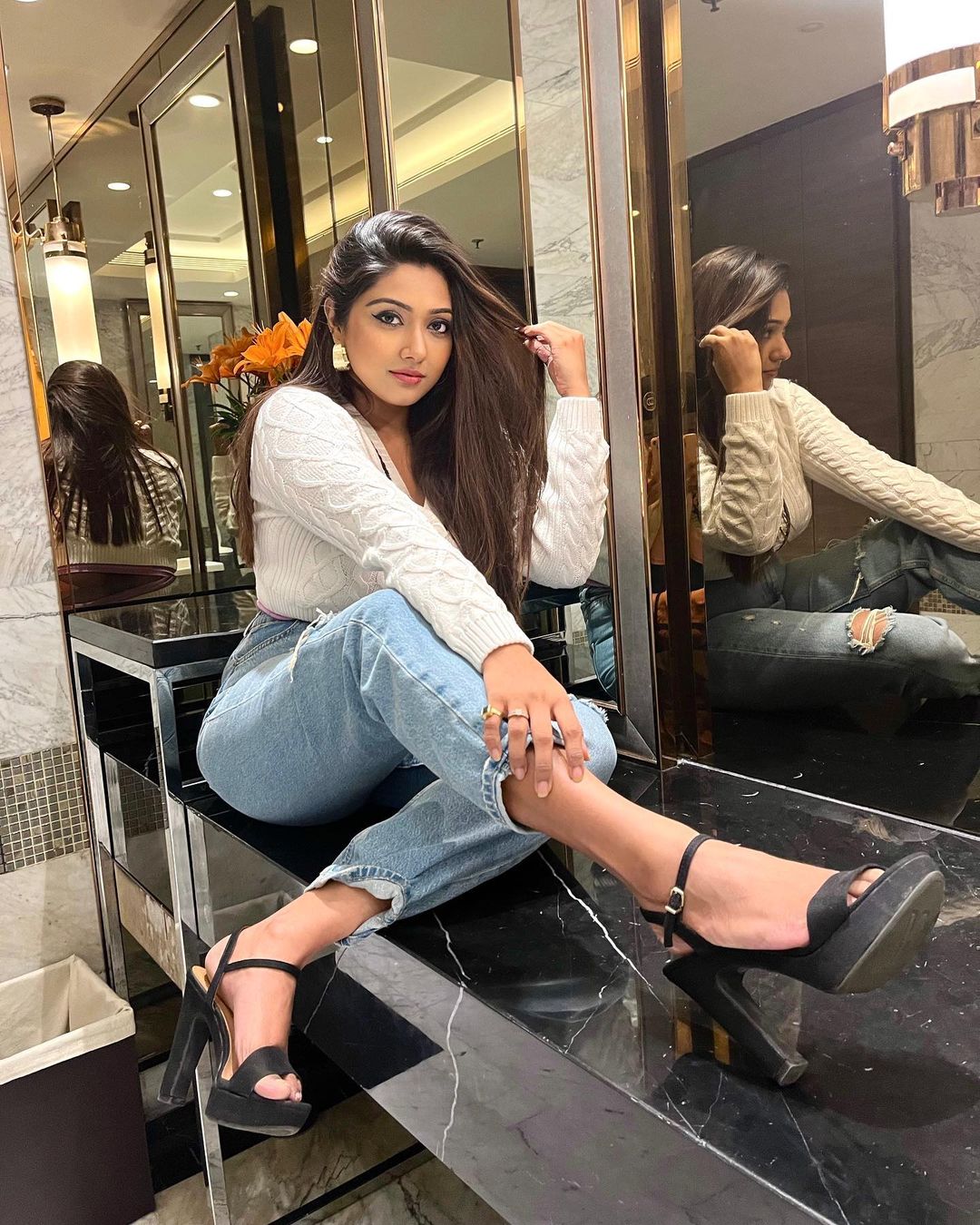 Agnijita Banerjee sitting on a black slab wearing a white sweater, blue jeans and black heels with her legs crossed and her left-hand holding her hair and right-hand rests over her right leg