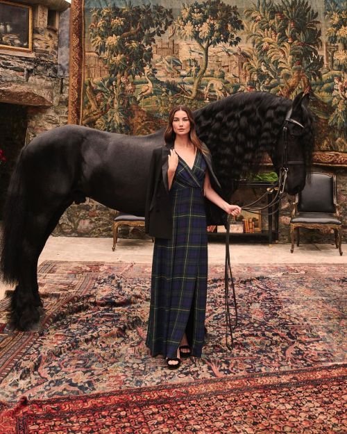 Glamorous photoshoot of Lily Aldridge in a black checks long-dress standing in front of a black horse holding the reins