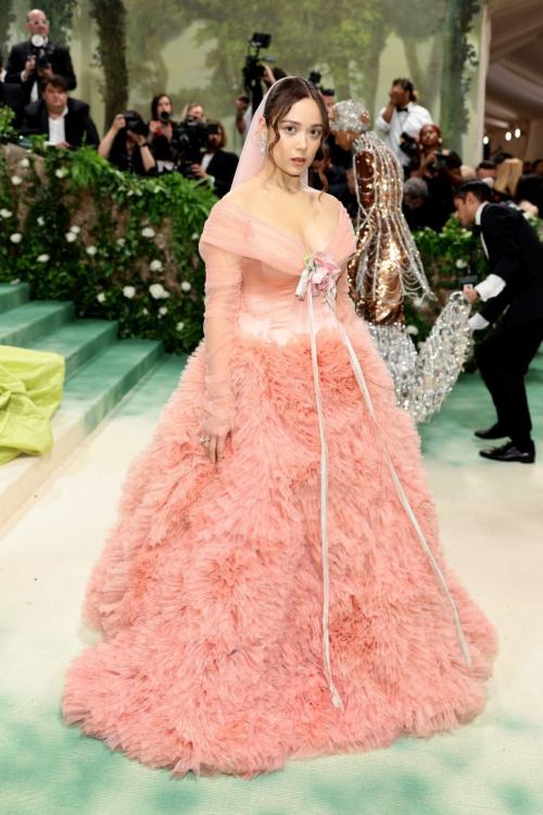Laufey posing at the 2024 Met Gala in a voluminous pink gown with a floral waist applique and sheer sleeves, channeling a Sleeping Beauty-inspired look.