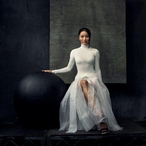 Anna Sawai in a white Dior turtleneck and sheer skirt seated gracefully against a minimalist dark backdrop.