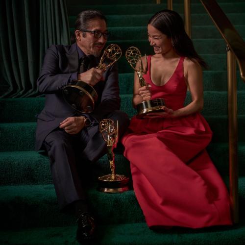 Anna Sawai and a fellow Emmy winner sharing a moment of laughter while holding their Emmy awards, seated on a green staircase.