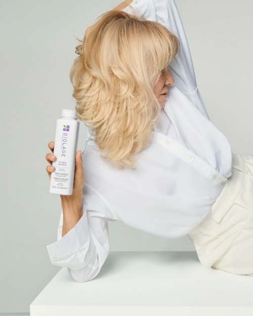 Back view of Pamela Anderson holding Biolage product while showcasing voluminous blonde layered hair against minimalist white background